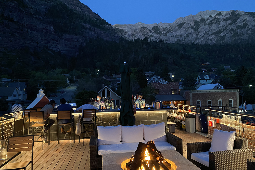 The Imogene rooftop bar at dusk, fire in the forefront, beautifully lit bar in the middle with the mountains in the background.