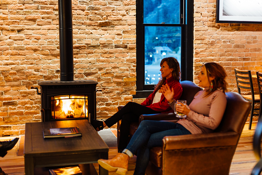 Image of the the Imogene's Whiskey bar seating area that is in front of the wood burning fire stove. There are people socializing in cozy leather chairs while enyoying a drink.