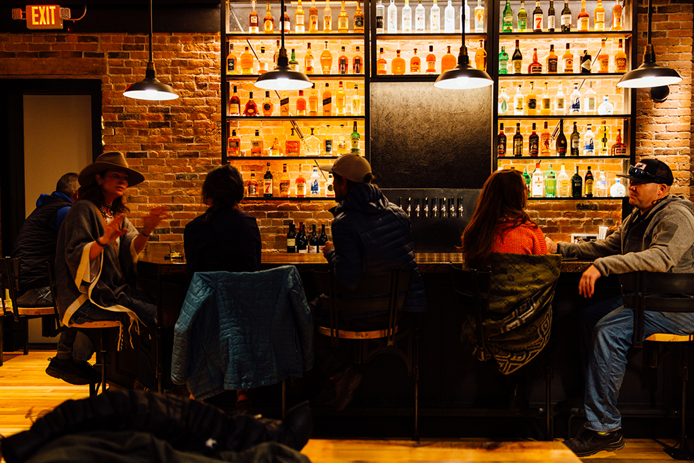 Image of the Imogene's whiskey bar, capturing the beautifully lit wall of bottles, and six people sitting on barstools socializing.